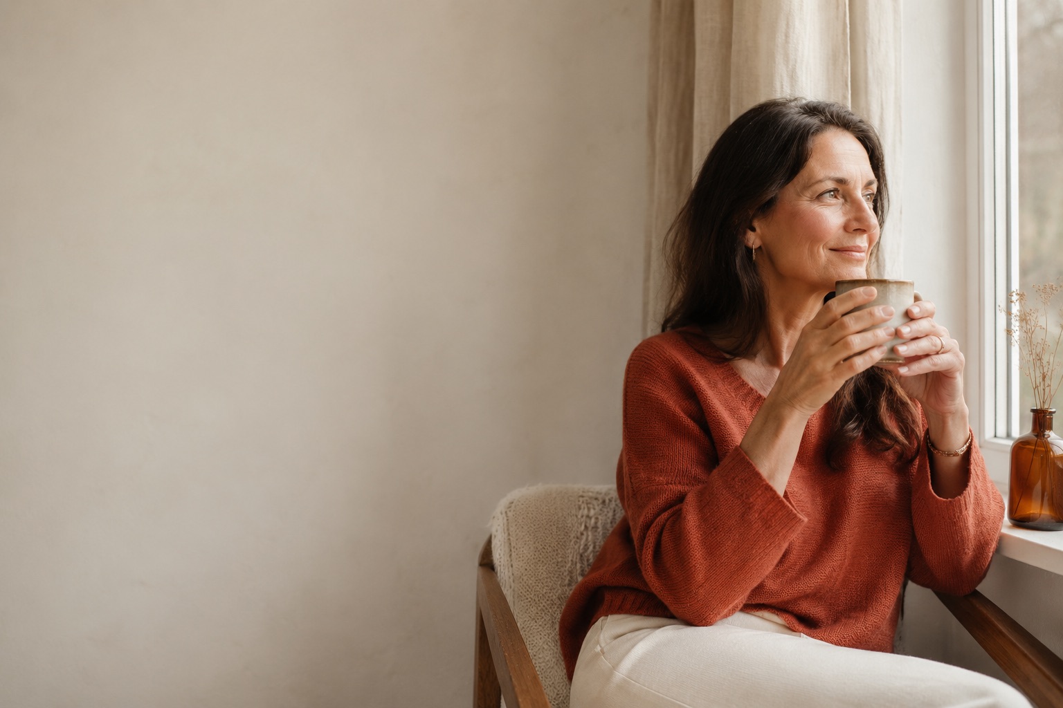Woman in a terracotta jumper sitting by a window holding a warm drink