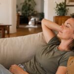 Woman lying back on a linen sofa with eyes closed, decompressing in warm light