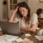 Woman at a cluttered kitchen table with sticky notes and a laptop, deep in thought