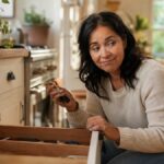 Woman crouched at an open kitchen drawer holding keys with a wry puzzled smile