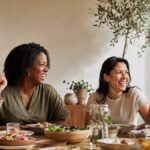 Four women friends laughing over a shared meal at a sunlit table