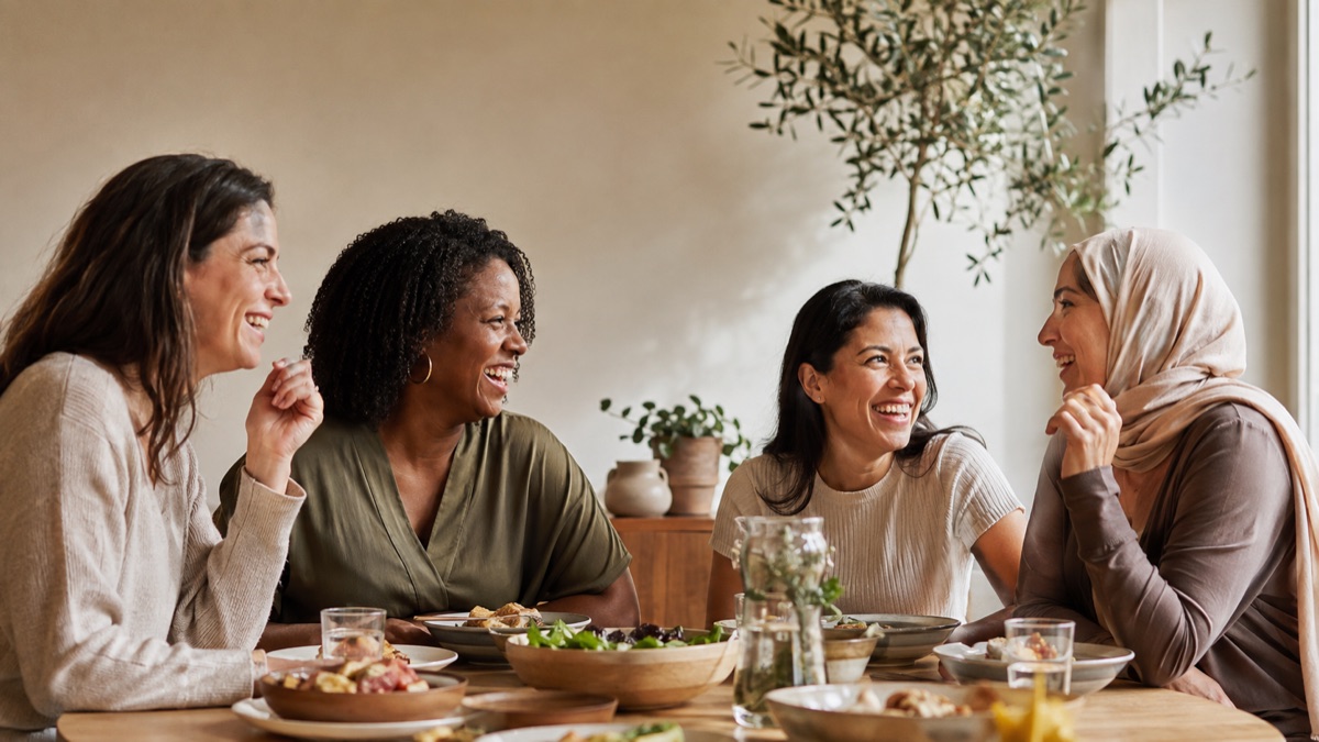 Four women friends laughing over a shared meal at a sunlit table