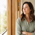 Woman in her forties by a window at home, arms crossed, looking out thoughtfully while considering menopause care options in Australia.