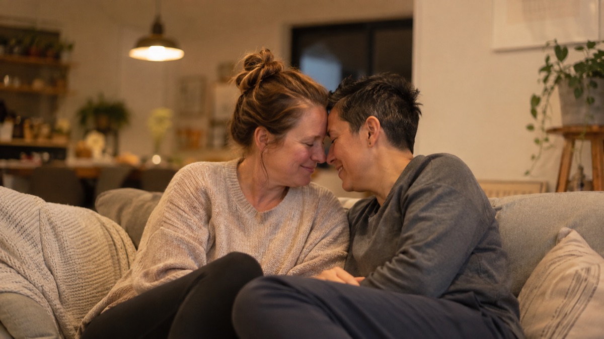 A couple sitting close on a sofa with foreheads gently touching in soft evening light