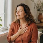 Woman around 47 with a hand on her chest taking a calming breath by a sunlit window, pausing during a busy day